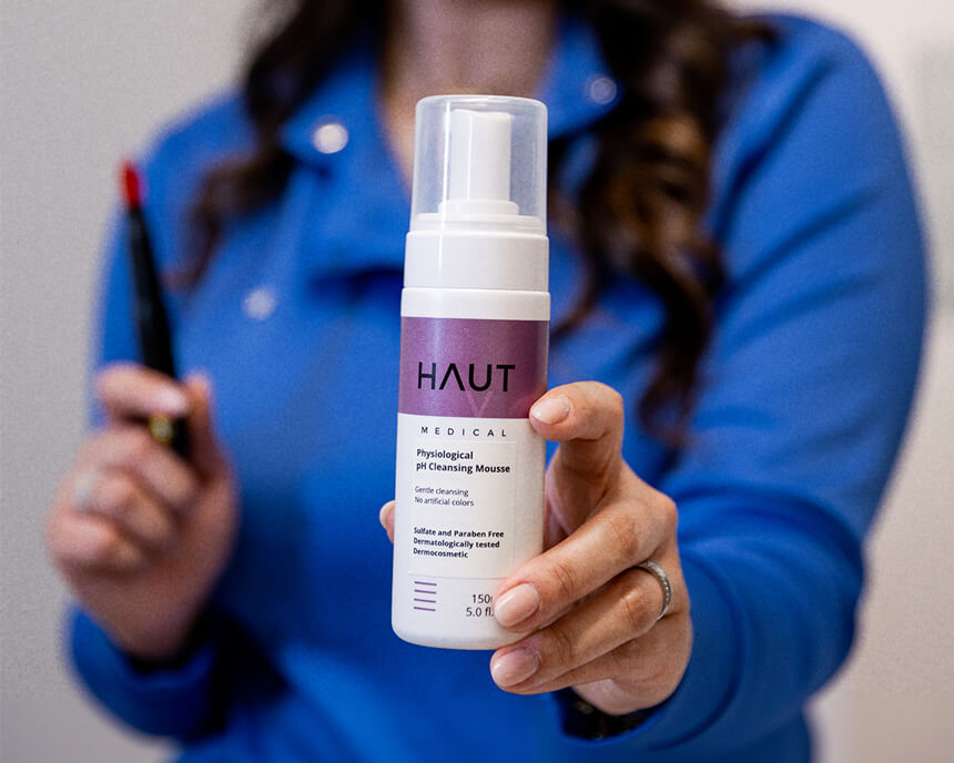 A beauty professional presents a bottle of HAUT MEDICAL Physiological pH Cleansing Mousse, highlighted against a blurred blue background.